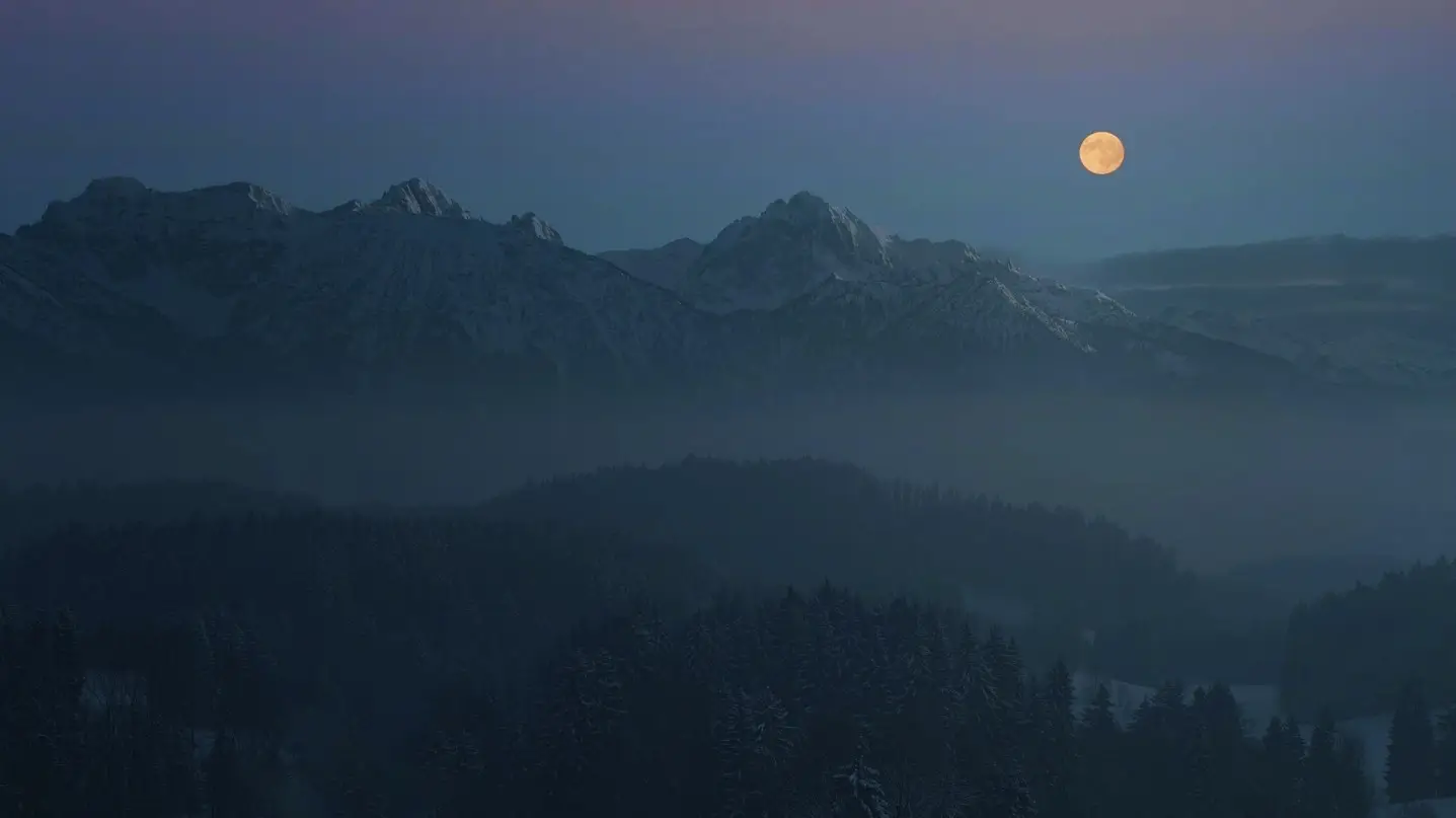 A full moon illuminates the mountains, casting a serene glow over the landscape in this captivating photograph.