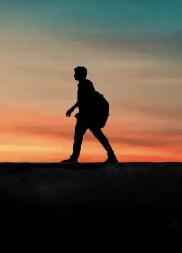 A silhouette of a man walking on a bridge, framed by the warm hues of a sunset in the background.